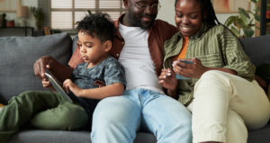 Young African American couple looking at screen of smartphone while sitting next to their son with touchpad watching online video