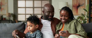 Young African American couple looking at screen of smartphone while sitting next to their son with touchpad watching online video